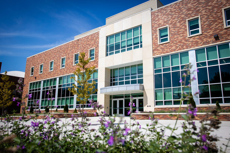 A modern university building with red brick and white exterior panels featuring multiple large windows under a clear blue sky, with purple flowering plants in the foreground and landscaped grounds at Northwest Missouri State University.